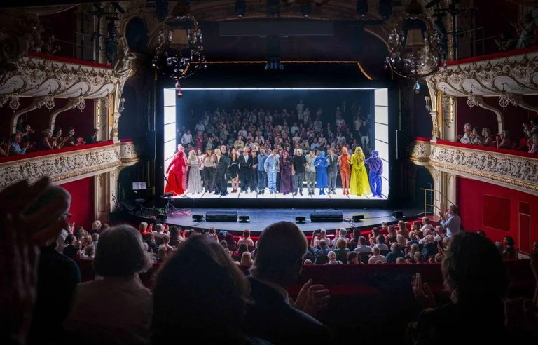 Theaterbühne mit Darstellern in bunten Kostümen verbeugt sich vor einem applaudierenden Publikum in einem geschmückten, traditionellen Theaterraum.