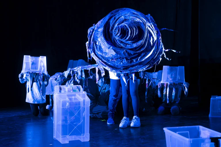 Personen in einem blauen Bühnenlicht tragen abstrakte Kostüme aus Kunststoff. Der zentrale Fokus ist eine große, spiralige Struktur.