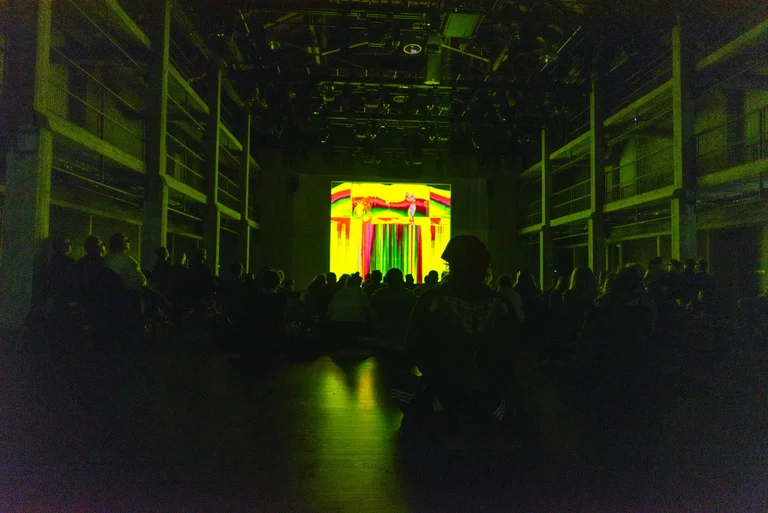 Dunkle Halle mit buntem, leuchtendem Bildschirm vorne. Menschen stehen oder sitzen, den Bildschirm beobachtend, möglicherweise bei einer Kunst- oder Multimedia-Show.