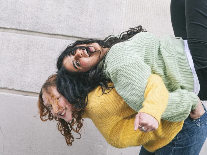 Zwei fröhliche Frauen in farbigen Pullovern lehnen sich rückwärts aneinander und lachen vor einem hellen Gebäudewand.