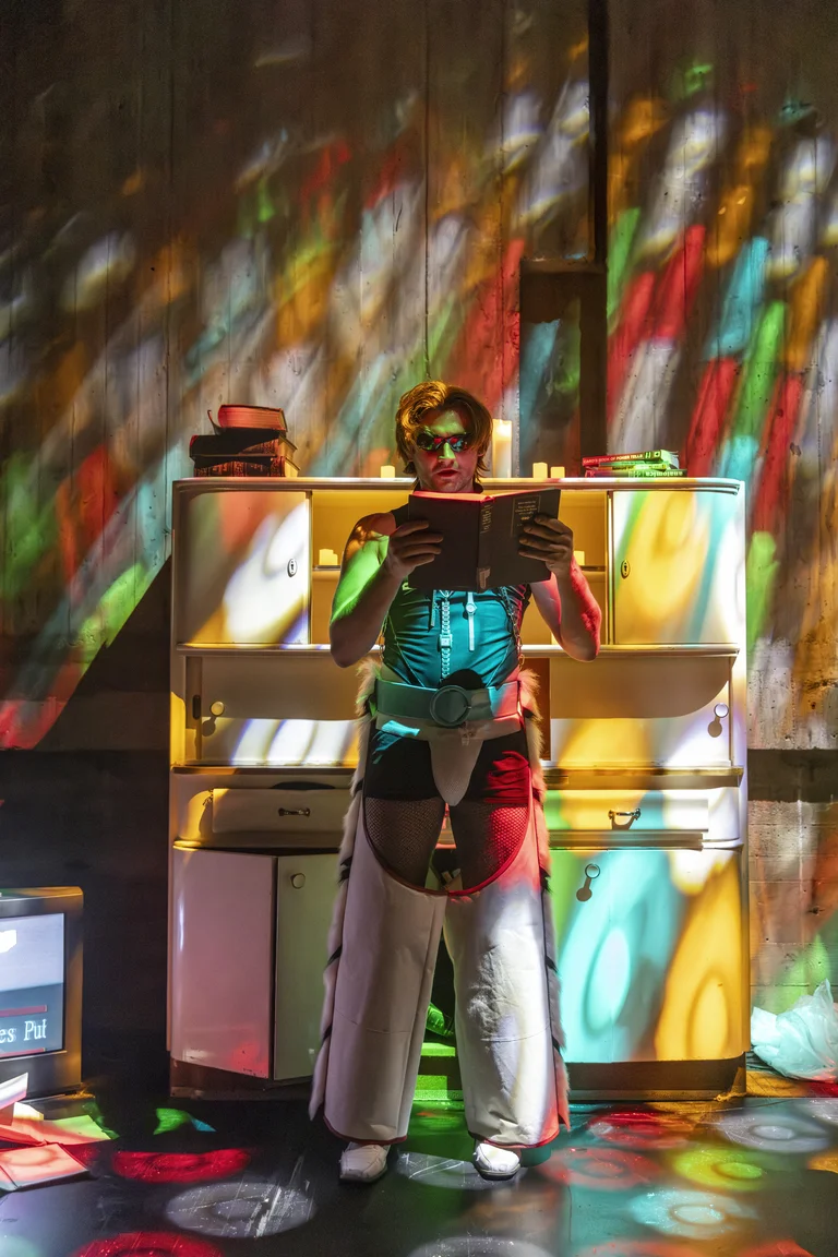 Eine Person in einem bunten futuristischen Kostüm liest ein Buch auf einer Bühne mit farbenfrohen Lichtprojektionen. Das Ambiente wirkt kreativ.