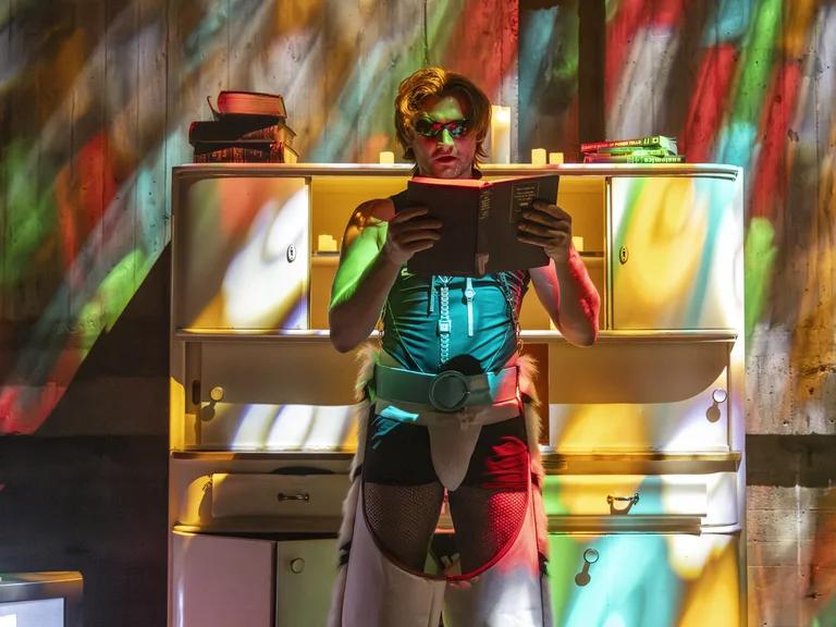 Eine Person in einem bunten futuristischen Kostüm liest ein Buch auf einer Bühne mit farbenfrohen Lichtprojektionen. Das Ambiente wirkt kreativ.