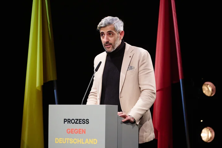 Ein Mann spricht auf einer Bühne mit einem Rednerpult, auf dem "Prozess gegen Deutschland" steht. Verschiedene farbige Fahnen im Hintergrund.