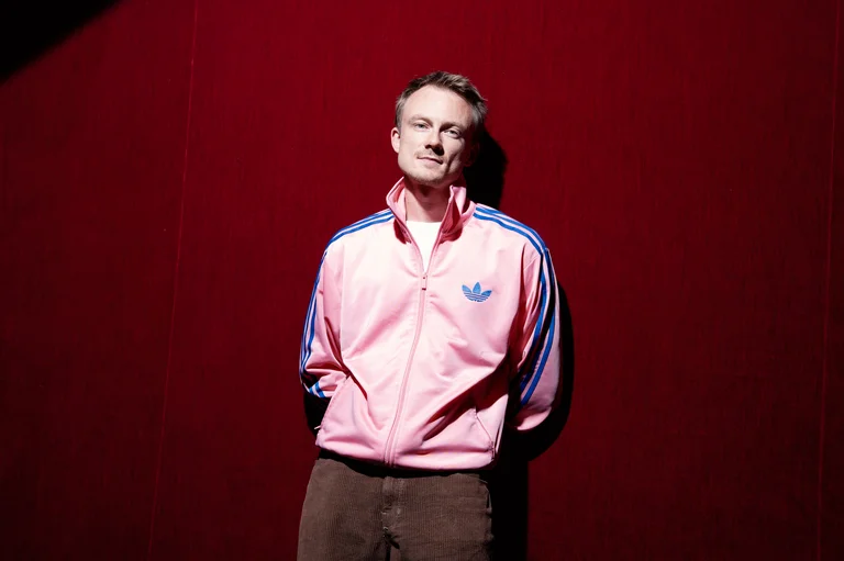 Person in einer rosa Adidas-Trainingsjacke steht vor einem tiefroten Hintergrund, schaut zur Kamera. Der Raum ist gut beleuchtet.