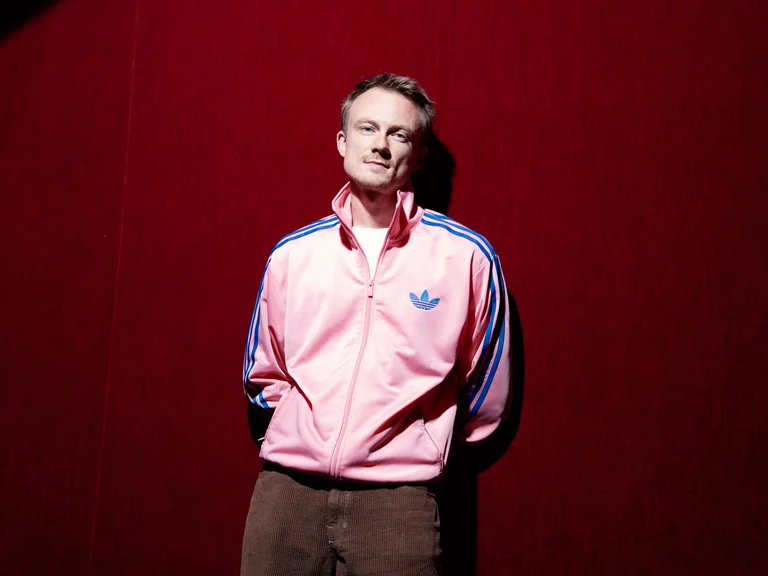 Person in einer rosa Adidas-Trainingsjacke steht vor einem tiefroten Hintergrund, schaut zur Kamera. Der Raum ist gut beleuchtet.
