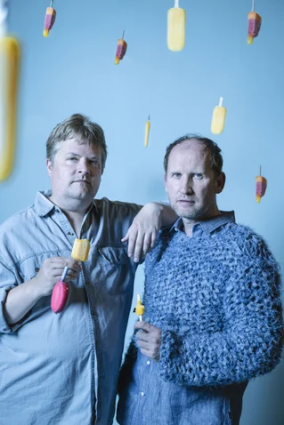 Two men pose with colorful popsicles. The background is blue with various floating popsicles in the air.