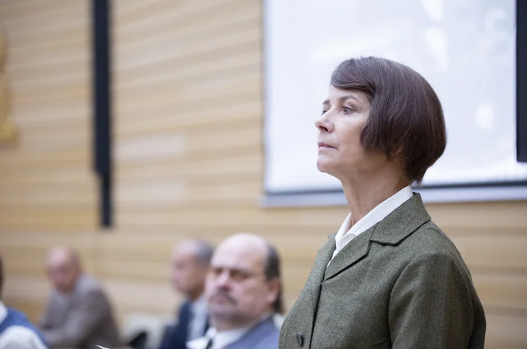 Eine Frau in einem Anzug steht in einem Konferenzraum, umgeben von anderen sitzenden Menschen. Sie schaut entschlossen.