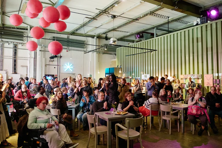 Eine große Gruppe von Menschen sitzt und steht in einem Veranstaltungsraum. An der Decke hängen rote Luftballons.