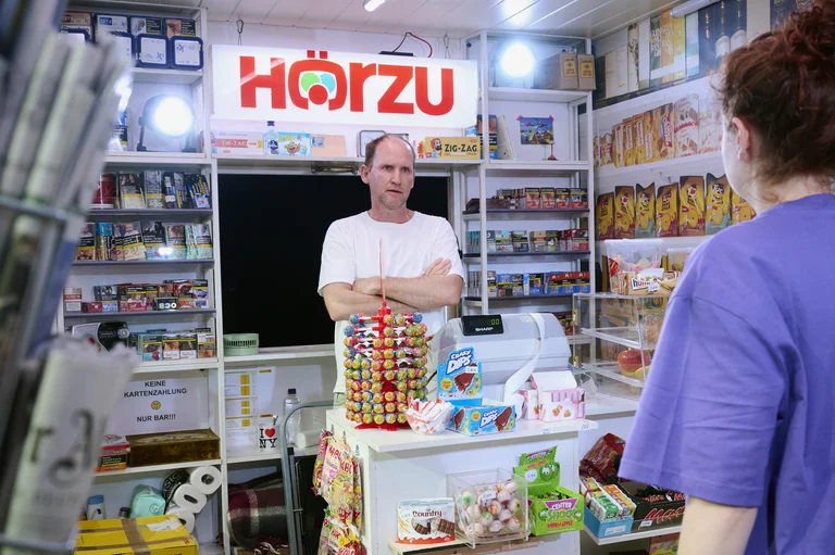Ein Mann steht hinter einem Kioskstand mit Zeitschriften, Süßigkeiten und Getränken. Ein anderes Gesicht zeigt Interesse am Stand.