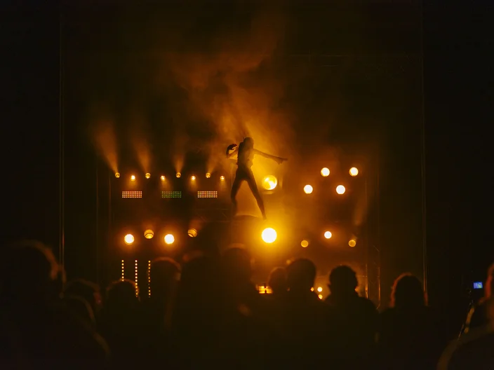 Ein Musiker tritt auf einer beleuchteten Bühne vor einem Publikum auf. Die Lichter sind warm, hauptsächlich in Orange- und Gelbtönen.