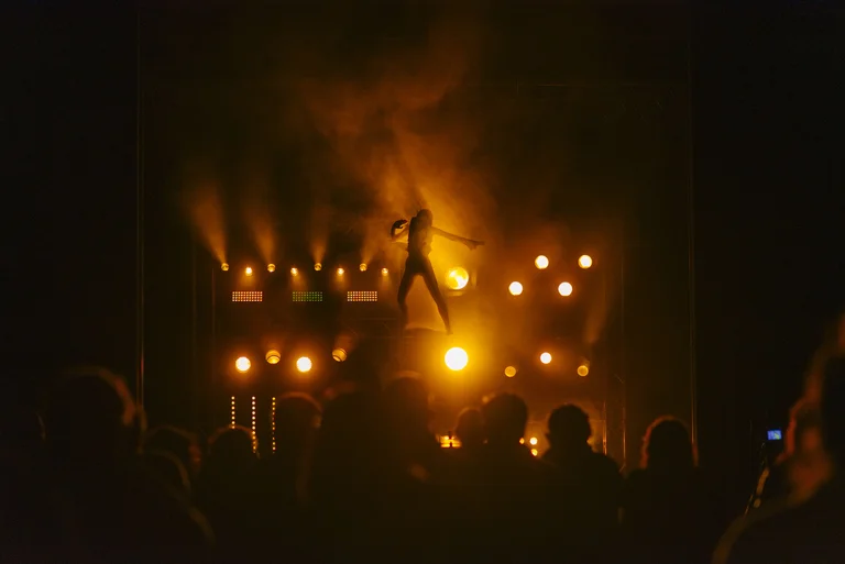 Ein Musiker tritt auf einer beleuchteten Bühne vor einem Publikum auf. Die Lichter sind warm, hauptsächlich in Orange- und Gelbtönen.