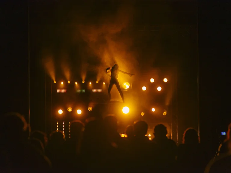 Ein Musiker tritt auf einer beleuchteten Bühne vor einem Publikum auf. Die Lichter sind warm, hauptsächlich in Orange- und Gelbtönen.