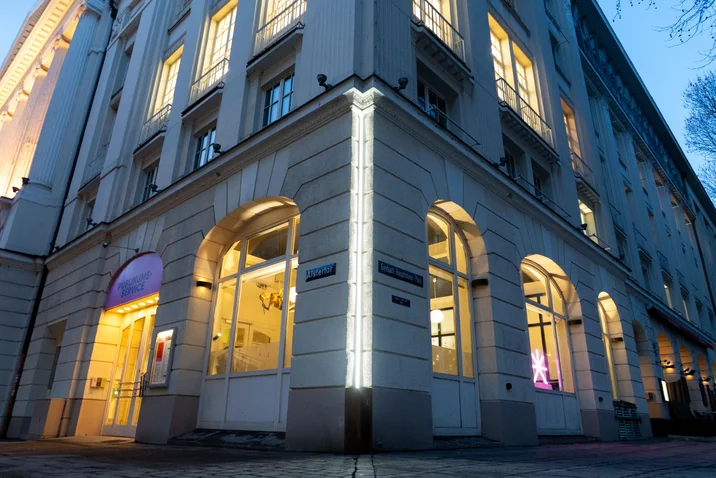 Außenansicht eines eleganten Gebäudes bei Nacht, beleuchtet mit gelbem Licht. Große Fenster und doppelte Eingänge auf jeder Seite.