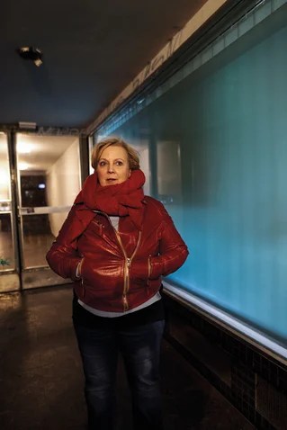 Eine Person steht in einem roten Mantel und Schal vor einem beleuchteten blauen Fenster in einem Flur.