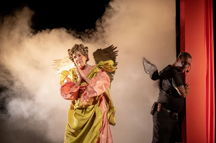 Two people on stage; one in colorful costume with wings, the other in black with wings; fog in the background.