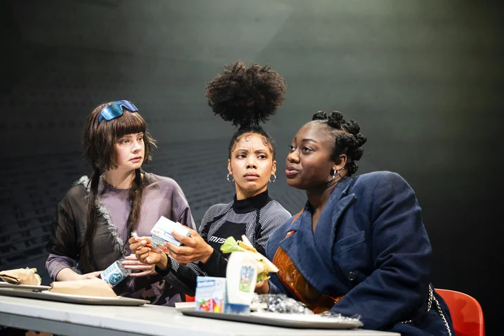 Three people sit at a table, engaged in discussion, with drinks and papers. Their clothing appears fashionable and unique.