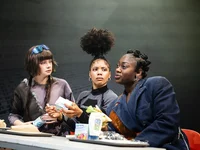 Three people sit at a table, engaged in discussion, with drinks and papers. Their clothing appears fashionable and unique.