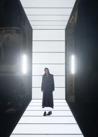 A person in a dark coat stands in the middle of a brightly lit runway, with shoes placed further away.