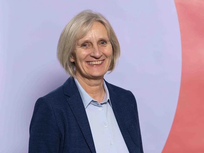 A smiling person with light-blonde hair wearing a dark blazer, standing against a pastel-colored background.