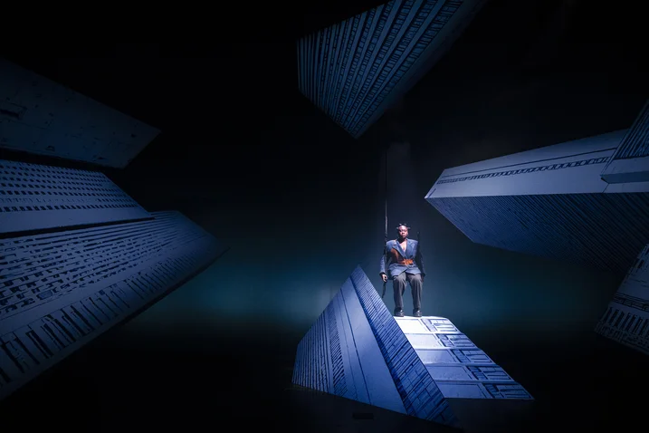 A person stands on a futuristic stage with glowing geometric forms, creating a dramatic and mysterious atmosphere.