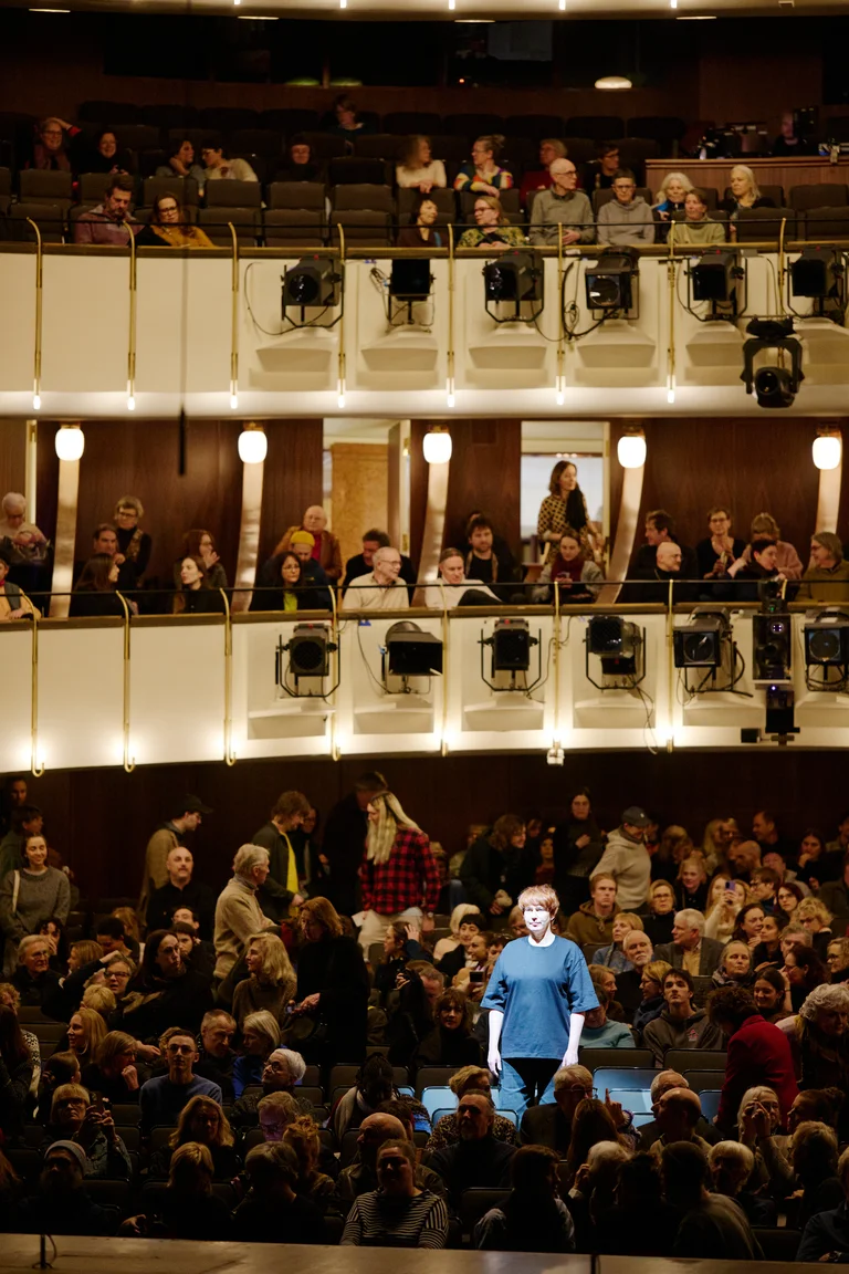 Eine große Menschenmenge sitzt in einem Theater. Eine Person steht im Vordergrund, von Scheinwerfern beleuchtet.