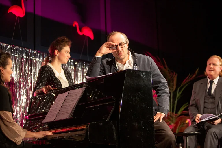 A group of people at a musical performance, featuring a pianist and two men in suits on a stage with flamingo decor.