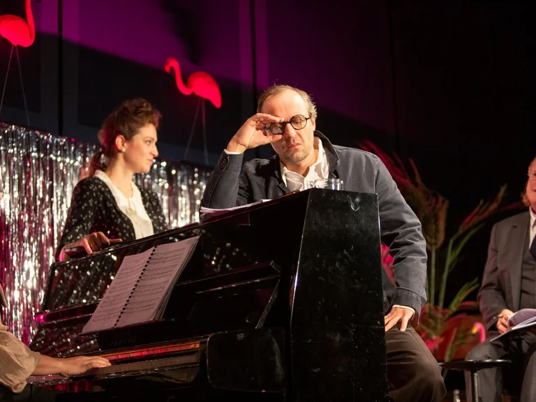 A group of people at a musical performance, featuring a pianist and two men in suits on a stage with flamingo decor.