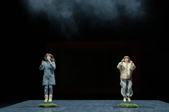 Two children in raincoats stand on small green platforms in a dark, misty stage setting, creating a mystical atmosphere.