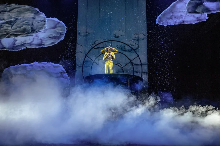 Ein Künstler in gelbem Outfit steht auf einer Bühne, umgeben von künstlichen Wolken und Nebel mit dramatischer Beleuchtung.