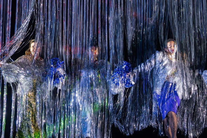 Three performers appear behind a curtain of shimmering, silver strands, as colorful lights illuminate the scene.