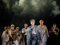 A group of actors in long coats performs on a foggy stage. The atmosphere is dramatic and mysterious.