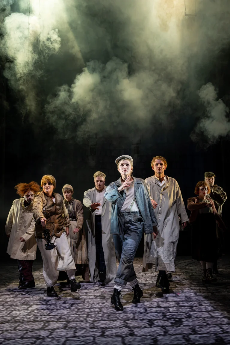 Eine Gruppe von Schauspielern in langen Mänteln tritt auf einer nebligen Bühne auf. Die Atmosphäre wirkt dramatisch und geheimnisvoll.