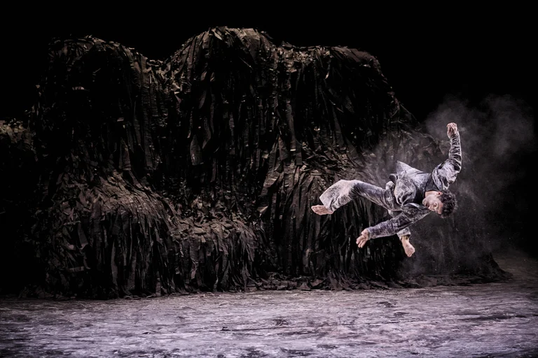 Ein Tänzer ist in einem dramatischen Sprung gegen einen dunklen, mystischen Hintergrund festgehalten. Bühne mit künstlerischem Staubeffekt.
