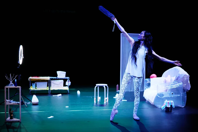 Eine Performerin steht auf einer Bühne voll beleuchtet, umgeben von Requisiten, in einer modernen Tanzvorstellung.