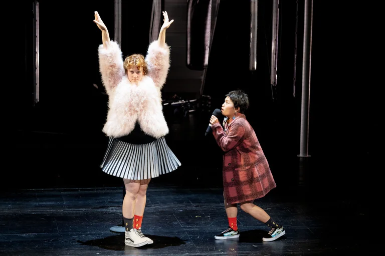 Zwei Performer auf einer Bühne. Eine Person trägt eine flauschige Jacke und hebt die Arme. Die andere singt.