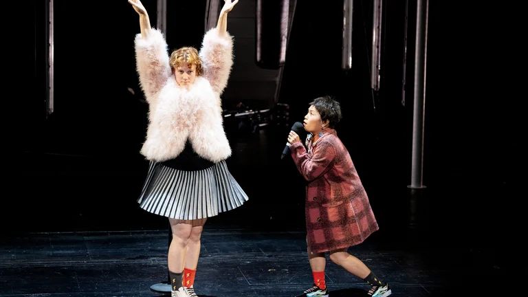 Zwei Performer auf einer Bühne. Eine Person trägt eine flauschige Jacke und hebt die Arme. Die andere singt.