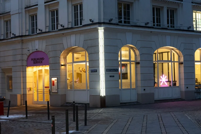 Ein beleuchtetes Thalia Theater bei Nacht, mit großen Fenstern und einer leuchtenden Skulptur im Inneren.