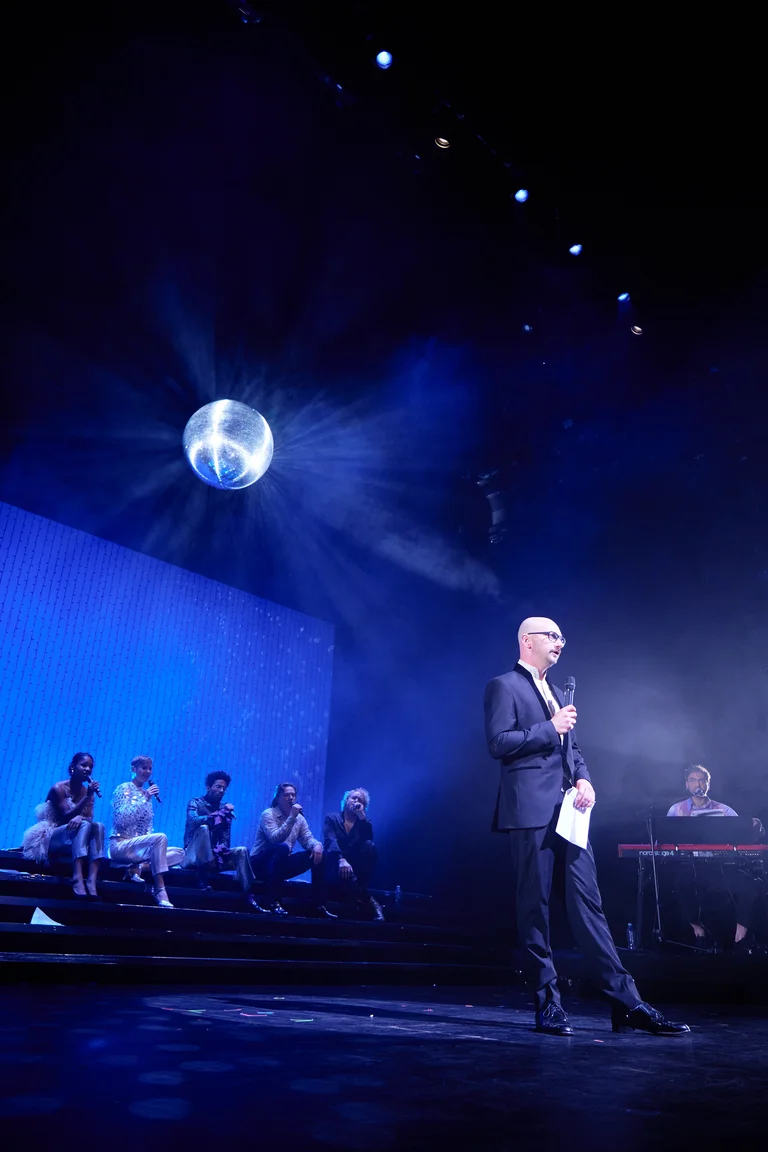 Eine Person in einem Anzug spricht auf einer Bühne. Im Hintergrund sind Musiker und ein glitzernder Ball zu sehen.