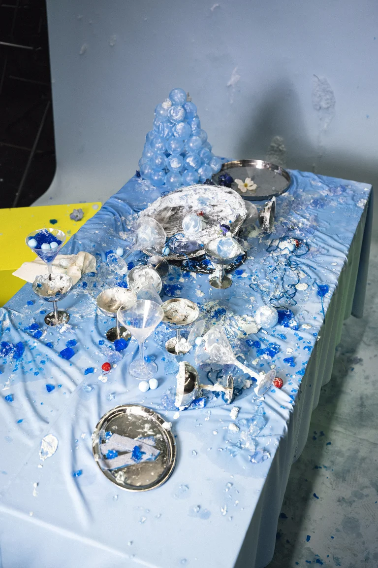 A table with a blue cloth is covered in spilled food and broken dishes, surrounded by scattered ice cubes.