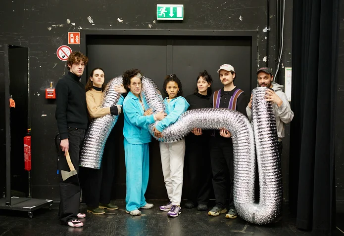 Eine Gruppe von sieben Personen in einem Raum hält ein großes, glänzendes silbernes Luftrohr und posiert vor einer schwarzen Tür.