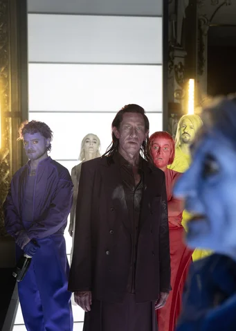 People stand in colorful suits with painted faces in a lineup. The background is illuminated with bright light.