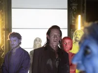 People stand in colorful suits with painted faces in a lineup. The background is illuminated with bright light.