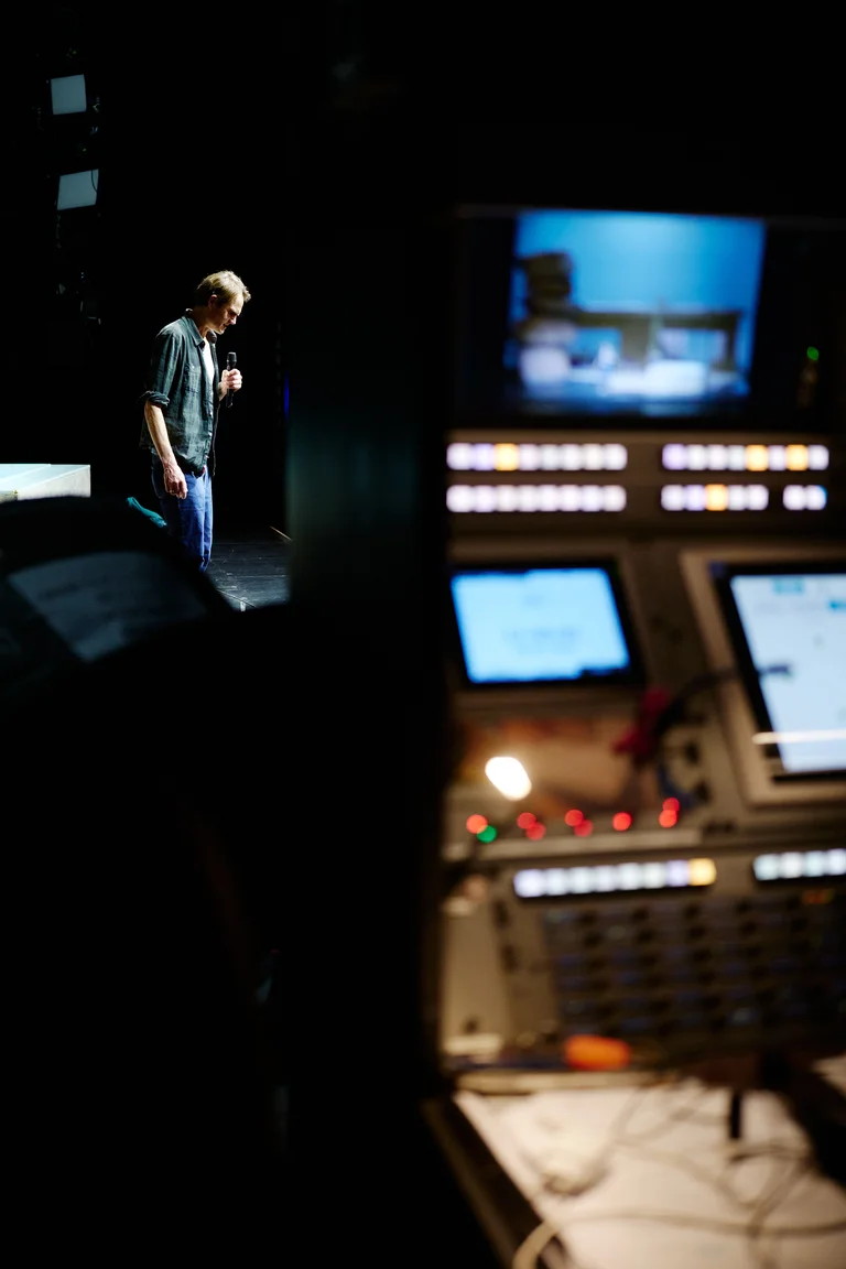 Ein Mann steht auf einer Bühne mit einem Mikrofon. Im Vordergrund sind unscharfe Lichter und Bildschirmanzeigen eines Mischpults zu sehen.