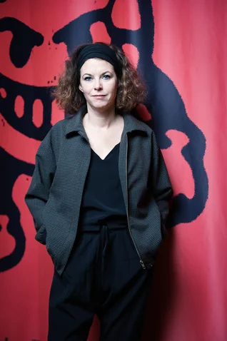 A woman with curly hair stands in front of a red backdrop with black patterns. She is wearing dark clothing and looks relaxed.