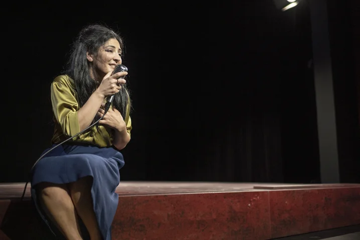 A woman sits on a stage holding a microphone. She appears happy and is speaking or singing.