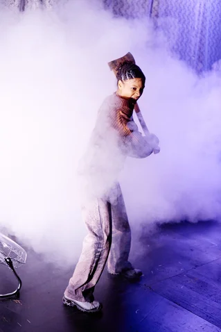 A performer with an axe stands in a dense cloud of fog on stage, with dramatic lighting enhancing the mysterious atmosphere.