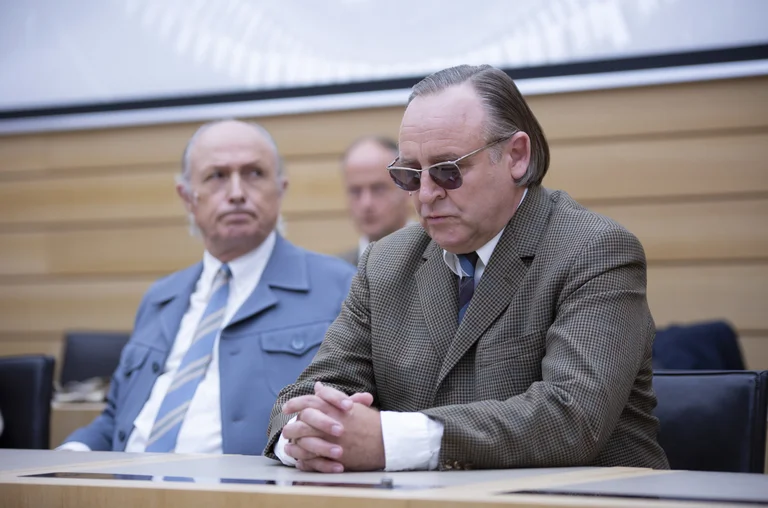 Zwei Männer sitzen in formeller Kleidung an einem Tisch. Einer trägt eine Sonnenbrille. Der Raum wirkt wie ein Konferenzsaal.