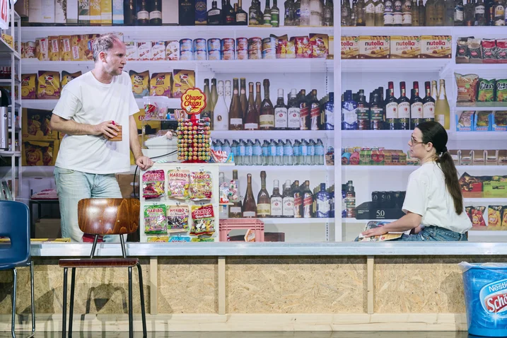 Two people perform on a stage set resembling a store. Shelves filled with products are visible in the background.