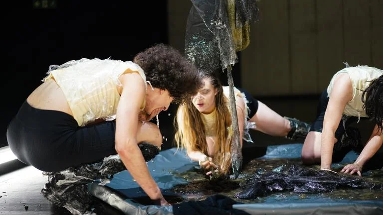 Drei Menschen in weißen Oberteilen und dunklen Hosen interagieren mit einem dunklen Stoff auf dem Boden bei einer Performance.