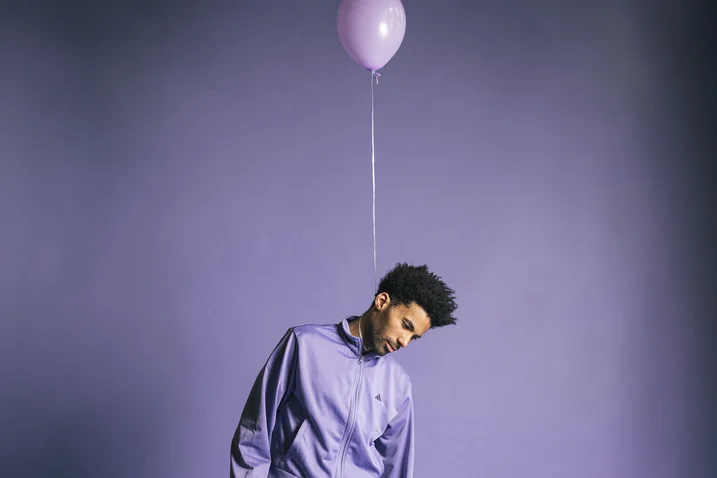 A man in a purple shirt tilts his head to the side, with a purple balloon floating above him.
